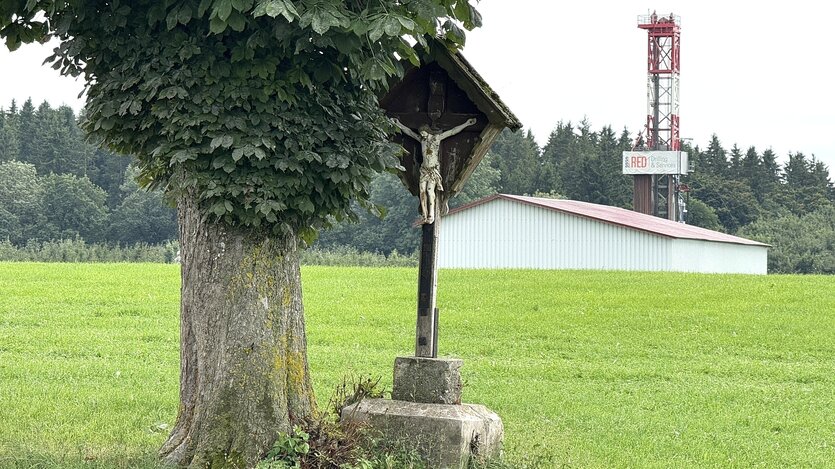 Ein Wegkreuz steht an einem Baum und im Hintergrund steht der Bohrturm, der untere Teil wird von einem Wellblechgebäude verdeckt