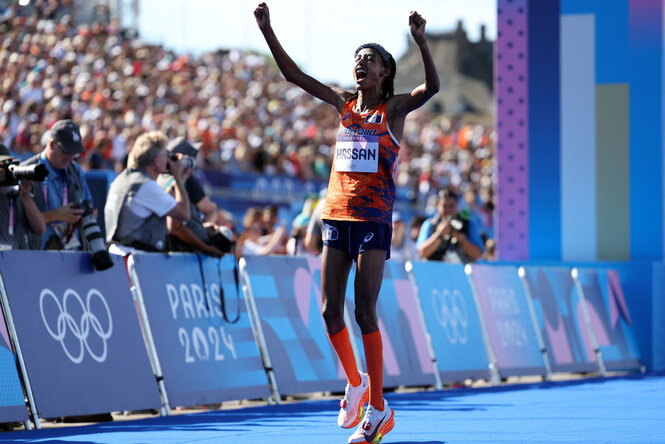 Das Bild zeigt die Läuferin Sifan Hassan im Moment des Siegesjubels bei den Olympischen Spielen in Paris 2024. Sie trägt ein orangefarbenes Trikot mit der Aufschrift „Nederland“ und dem Namensschild „Hassan“. Im Hintergrund ist eine große Menschenmenge sowie Fotograf:innen zu sehen Links und rechts sind die olympischen Ringe und das Logo „Paris 2024“ sichtbar.