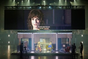 Zwei Kameraleute filmen in einem Bühnenset mit drei Räumen, darüber ist eine Frau im Video projiziert