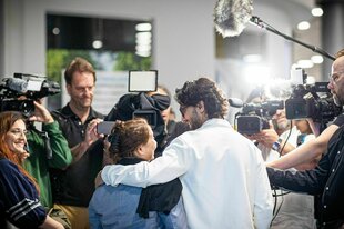 Zwei Personen stehen Arm in Arm vor einer Gruppe von Journalisten