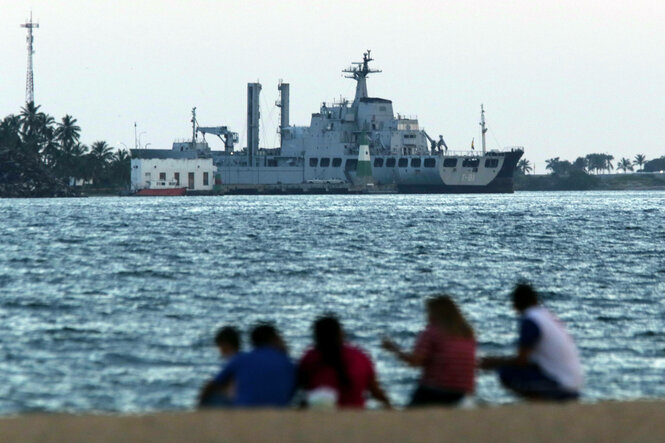 Im Vorderdrund sitzen Menschen am Wasser, im Hintergrund ist ein Kriegsschiff zu sehen