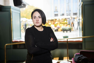 Eine Frau steht mit verschrenkten Armen vor einem Fenster
