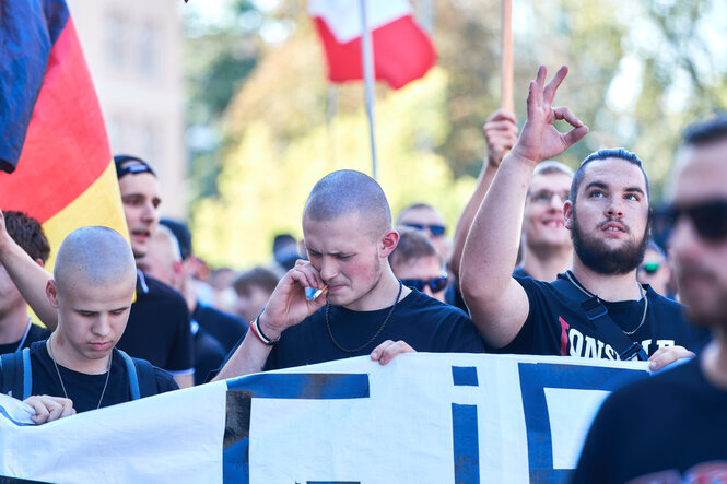 „Gruppe junger Männer nimmt an einer Demonstration teil, einige halten ein Transparent. Im Hintergrund sind Nationalflaggen zu sehen. Eine Person macht mit der Hand ein Symbol, die Finger formen ein W und P, vermutlich ist damit der rechtsexttreme Ausdruck „White Power“ gemeint. Die Stimmung wirkt angespannt und konzentriert.“