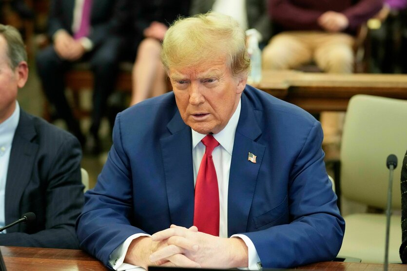 Donald Trump trägt einen blauen Anzug, ein weißes Hemd und eine rote Krawatte. Er sitzt in einem Gericht am Tisch , guckt ernst und hat die Hände vor sich auf dem Tisch gefaltet.