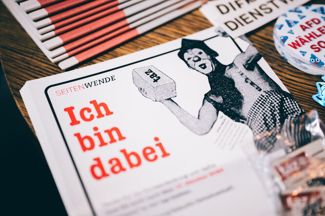 Das Bild zeigt einen Flyer, auf dem „Ich bin dabei“ steht. Neben dem Schriftzug ist eine Person zu sehen, die einen Stein in die Höhe hält, auf dem taz steht.