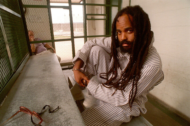 Der Journalist Mumia Abu Jamal sitzt im Schneidersitz in weißen Kleidern in einer Gefängniszelle