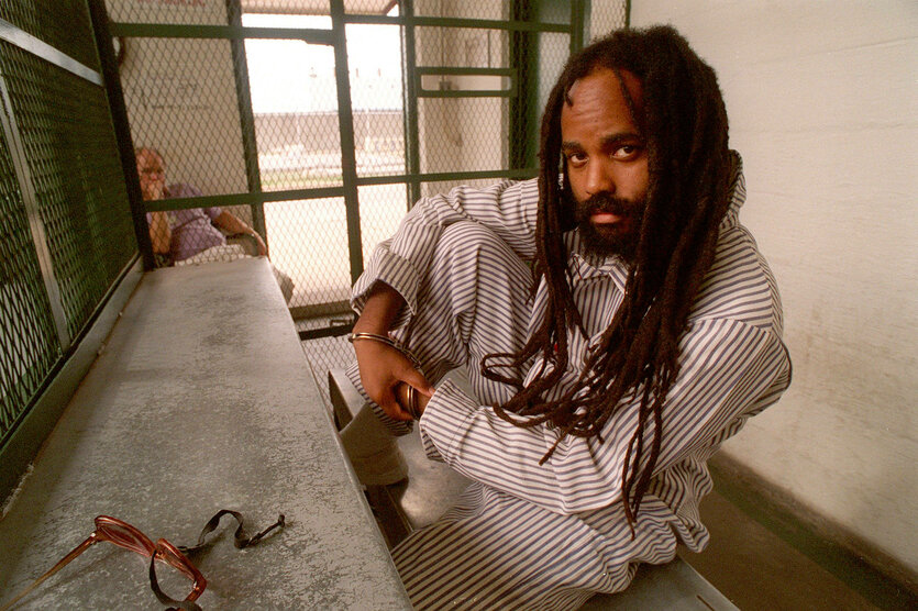 Der Journalist Mumia Abu Jamal sitzt im Schneidersitz in weißen Kleidern in einer Gefängniszelle