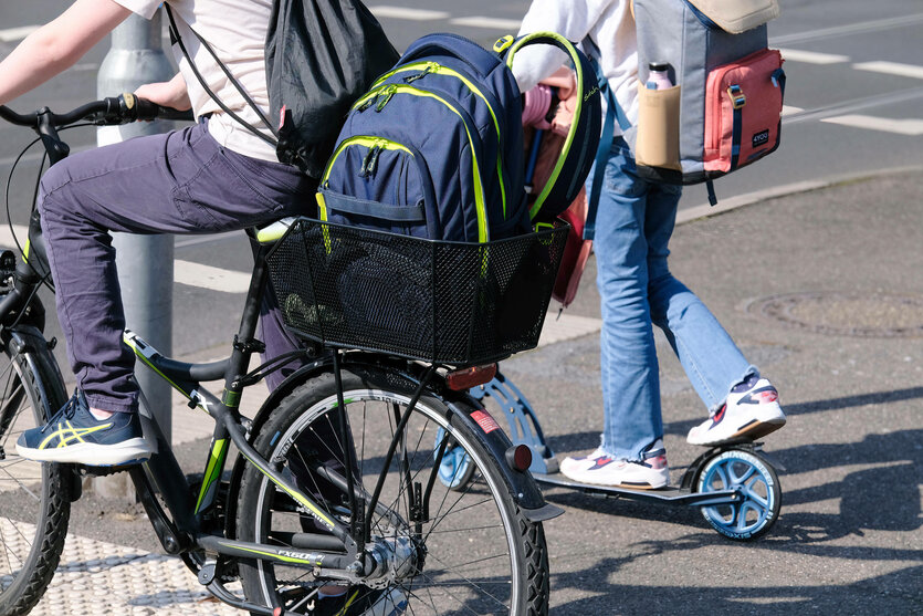 Zwei Schulkinder auf dem Schulweg, eines unterwegs mit dem Fahrrad, das andere mit dem Tretroller