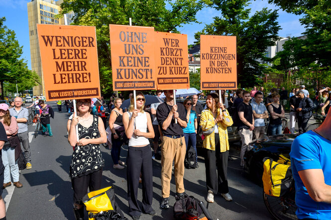 Angestellte der Universität der Künste Berlin stehen auf einer Straße und protestieren, sie halten orange Schilder mit Aufschriften wie "Weniger Leere, mehr Lehre" oder "Wer kürzt, denkt zu kurz"