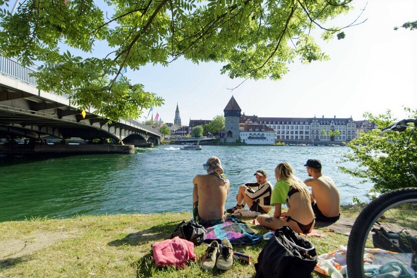 Junge Menschen in Badekleidung sitzen am Ufer eines Sees.
