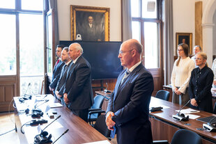 Das Bild zeigt einen Gerichtssaal mit Personen in Anzügen vor einem Tisch mit Mikrofonen und Dokumenten. Im Vordergrund steht ein Mann mit verschränkten Händen.