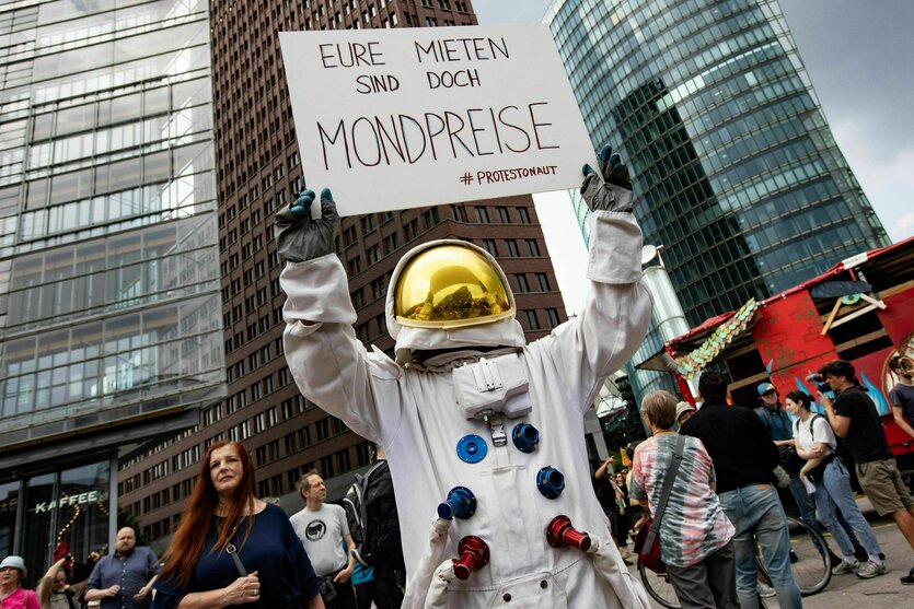 Bei einer Demo gegen hohe Mieten hält ein als Astronaut verkleideter Demonstrant am Potsdamer Platz vor Hochhäusern ein Schild hoch. Auf dem steht: "Eure Mieten sind doch Mondpreise"