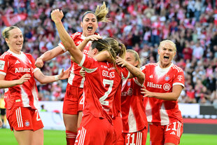 Junge Frauen in FC-Bayern-Trikots jubeln auf dem Rasen, hinter ihnen eine volle Stadiontribüne