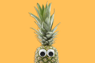 Eine Ananas vor dunkelgelbem Grund. Die Ananasfrucht ist erst ab der oberen Hälfte im Bild. Darauf geklebt sind zwei Spielzeugaugen. Viel größer als die Frucht: die grüne Krone, die bis an den oberen Bildrand reicht.