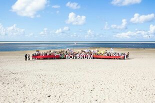 Ein Strandbild mit Menschen, die ein rotes Banner mit der Aufschrift "BORKUM" halten, unter einem blauen Himmel mit Wolken.