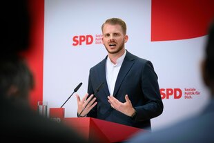 Tim Klüssendorf steht vor einer Wand mit dem SPD Logo und spricht in ein Mikrofon
