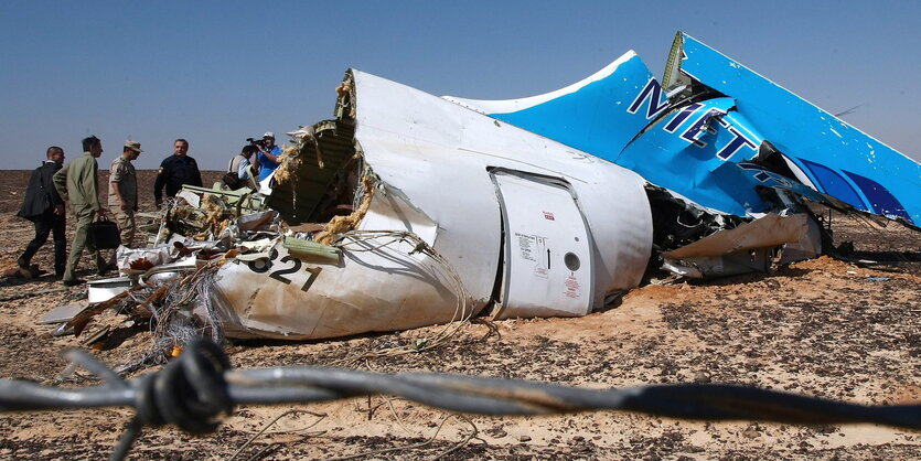 Wrackteile des in Ägypten abgeschossenen russischen Flugzeugs. Im Hintergrund sind ein paar Menschen vor blauem Himmel zu sehen, die die Absturzstelle untersuchen. Vor der Kameralinse verläuft unscharf Stacheldrahtzaun.