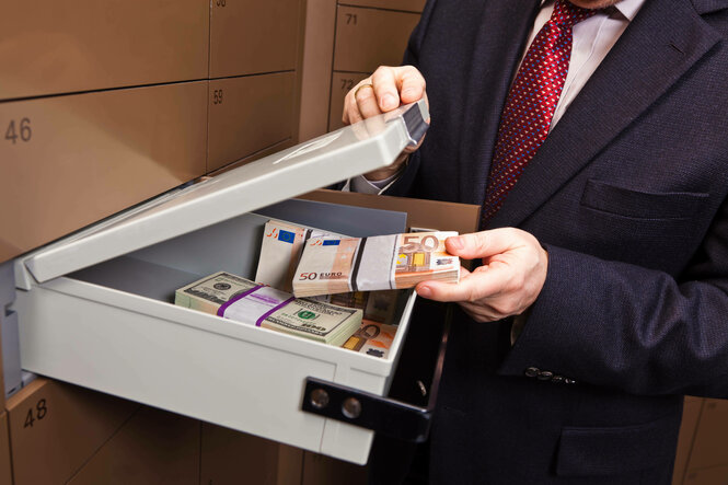 Ein Mann mit Krawatte öffnet ein Schliessfachschublade, in dem sich Bündel von Banknoten befinden