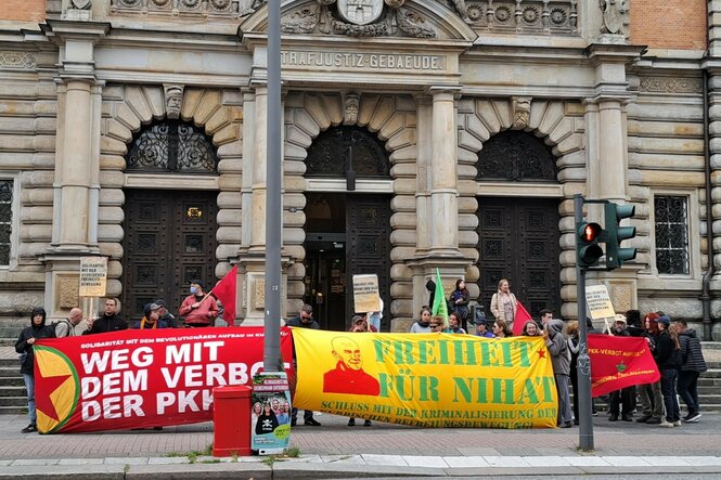 Vor dem Strafjustizgebäude stehen Aktivist:innen mit drei Bannern und Plakaten. Auf den Bannern steht Freiheit für Nihat, Weg mit dem Verbot der PKK und Freiheit fü alle politischen Gefangenen.