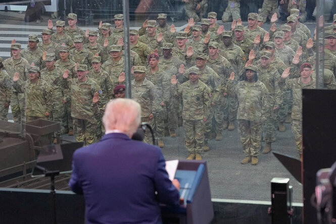 Donald Trump steht hinter einer Glasscheibe und spricht zu US-Soldaten in Uniform die salutieren