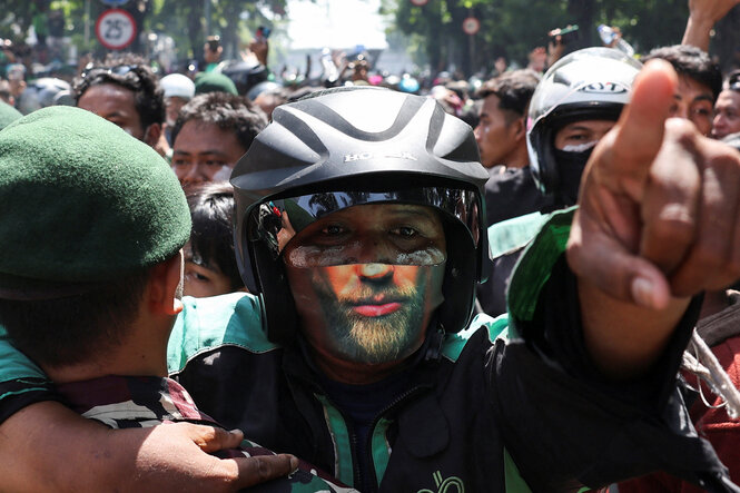 Ein Demonstrant mit Helm und bemaltem Geischt steht in einer Menschenmenge, sein Arm ist nach vorne gestreckt