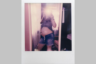 Eine junge schwangere Frau in kurzen Jeans-Hosen steht vor dem Spiegel und macht ein Selfie von sich
