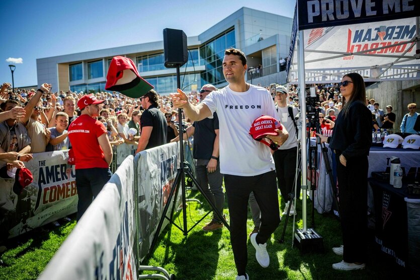 Charlie Kirk verteilt Hüte, er trägt ein weisses T-Shirt mit der aufschrift "Freedom" - hinter einer Absperrung stehen junge Menschen, die einen der Hüte fangen wollen