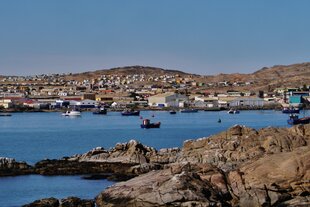 Blick von Shark Island auf die Küste der namibischen Stadt Lüderitz. Im Vordergrund liegt eine felsige Küste, dahinter eine ruhige Bucht mit mehreren kleinen Fischerbooten auf dem Wasser. Am gegenüberliegenden Ufer erstreckt sich Lüderitz mit vielen bunt gestrichenen Häusern, die sich über einen sanft ansteigenden Hügel verteilen. Der Himmel ist klar und blau , ein sonniger Tag im Atlantik.