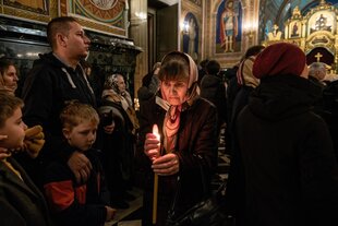 Gläubige stehen dicht gedrängt in einer Orthodoxen Kirche, eine ältere Frau trägt eine brennende Kerze