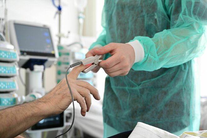 Ein Pfleger in grüner Schutzkleidung legt in einem Krankenzimmer bei einem Patienten ein Messgerät am Finger an