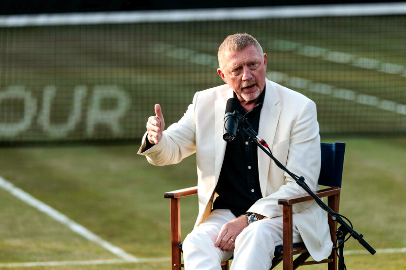 Boris Becker sitzt auf einem Rasentennisplatz auf dem Stuhl, gestikuliert und spricht in ein Mikrofon