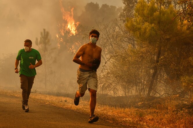 Zwei Menschen mit kurzen Haaren, der eine oberkörperfrei mit kurzer Hose, der andere mit langer Hose und grünem T-Shirt, rennen durch eine trockene Landschaft. Hinter ihnen stehen Bäume in Flammen, die Lust ist voller Rauch und vom Feuer orange gefärbt. Beide Menschen tragen Atemschutzmasken.