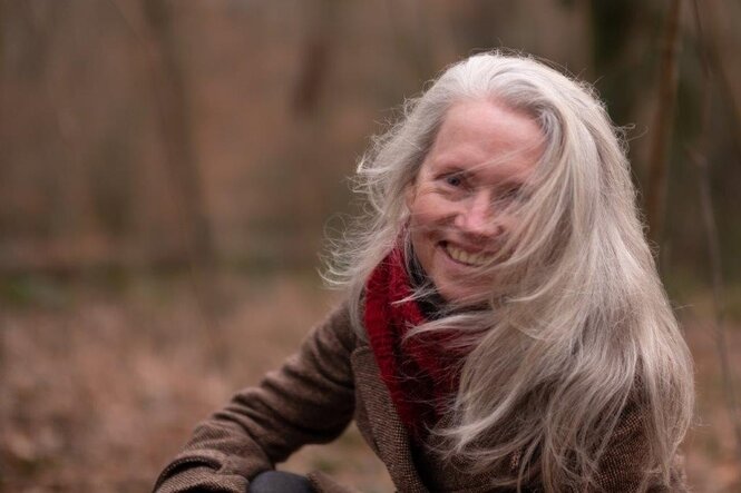 Graues langes Haar weht einer Person ins Gesicht und bedeckt dieses zur Hälfte