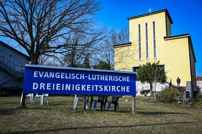 Vor einem gelben modernen Kirchengebäude steht auf einem Schild "Evangelisch-lutherische Dreifaltigkeitskirche. Auf einer Treppe vor der Kirche geht ein Mann mit schwarzen Haaten