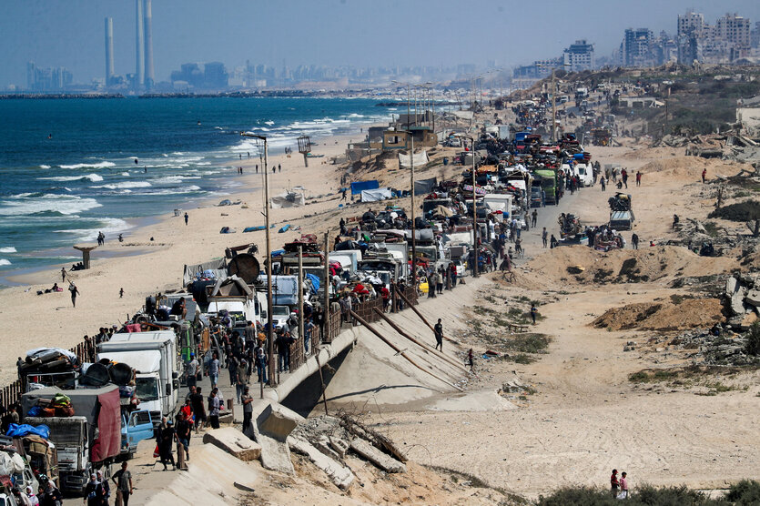 Eine große Wagenkolonne, die von Fußgängern begleitet wird, fährt den Strand entlang. Links das Meer und der Strand