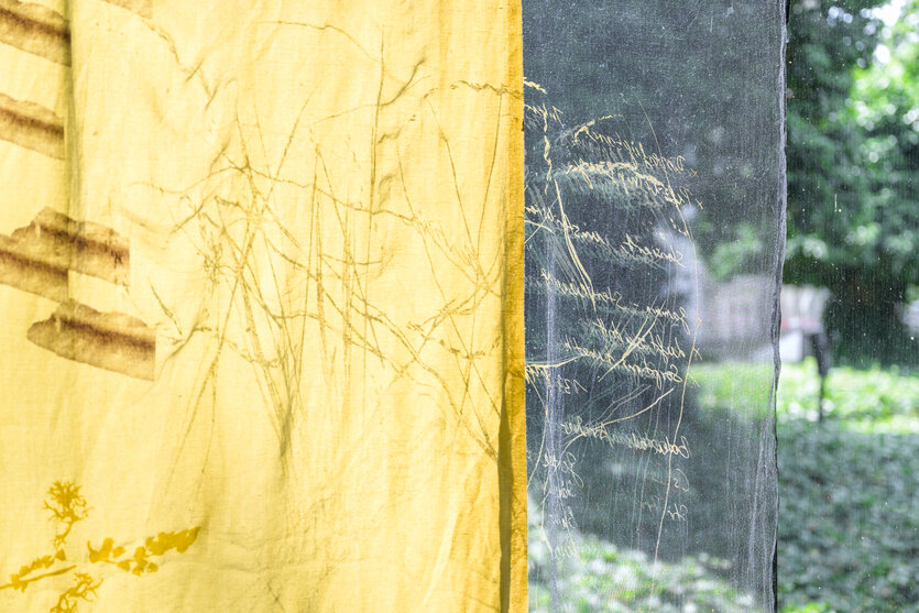 Eine Stoffbahn mit Pflanzenformen, im Hintergrund der Blick durch ein Fenster auf Bäume.