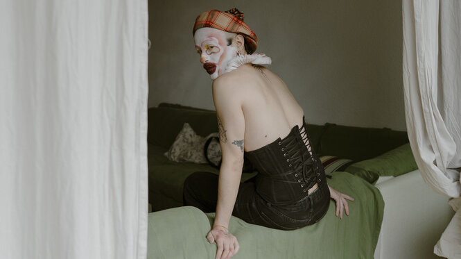 Fagatta sitzt auf dem Rand eines Sofas, links und rechts hängen helle Vorhänge. Das Gesicht ist clownesk geschmickt, er trägt eine Mütze und ein Korsett