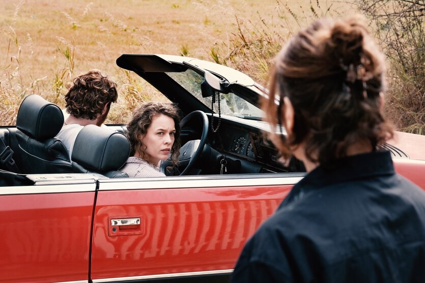 Paula Beer als Beifahrerin in einem roten Cabriolet in der Uckermark, im Hintergrund ein Feld