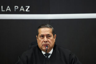 Mann in Anzug und Taler, mit zweifelendem BLick vor schwarzer Wand auf der oben links noch "La Paz" lesbar ist