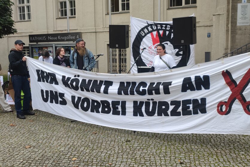 Demonstrant:innen stehen vor dem Rathaus in Treptow mit einem Transparent. Darauf steht: Ihr könnt nicht an uns vorbei kürzen.