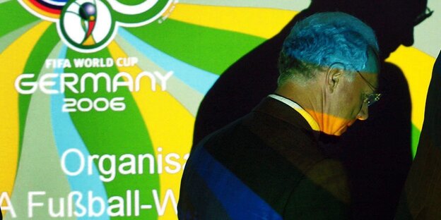 Franz Beckenbauer steht neben einem Plakat zur WM 2006. Sein Kopf wirft einen dunklen Schatten auf das Plakat.