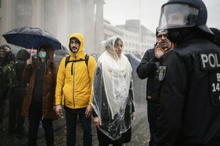 Menschen stehen in Regenkleidung zusammen vor dem Brandenburger Tor in Berlin, sie schauen finster, ein Polizist steht am Bildrand