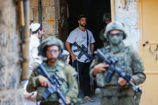Ein mit einem Maschinengewehr bewaffneter Siedler steht in einem Hauseingang, vor ihm stehen zwei schwer bewaffnete israelische Soldaten zu sehen