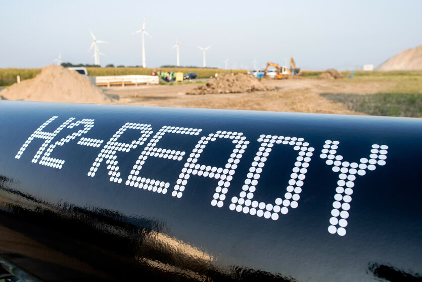 Ein großes Transparent mit der Aufschrift H2-Ready (also ungefähr: bereit für Wasserstoff) steht vor einer kargen Sandfläche mit Bagger; im Hintergrund stehen Windräder