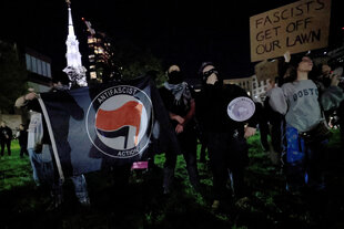 Menschen stehen nachts draußen, sie halten eine Fahne hoch, auf der "antifacist action" steht, und ein Plakat mit der Aufschrift "Facist get off our lawn"