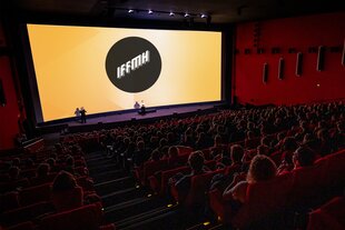 Großer, moderner Kinosaal mit roten Sitzen, viele Menschen blicken auf eine große Leinwand, auf der das Logo des Internationalen Filmfestivals Mannheim-Heidelberg zu sehen ist. Vorne stehen zwei Personen auf der Bühne.
