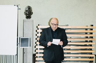 Zu sehen ist der Soziologe Armin Nassehi der einen Zettelblock schaut. Im Hintergrund eine Seminarraum Umgebung, mit gestapelten Tischen, einem Whiteboard, einer Büste und einem Pult.