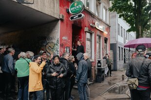 Menschen stehen vor einem Haus mit roter Fassade, Graffiti und Bierwerbung.