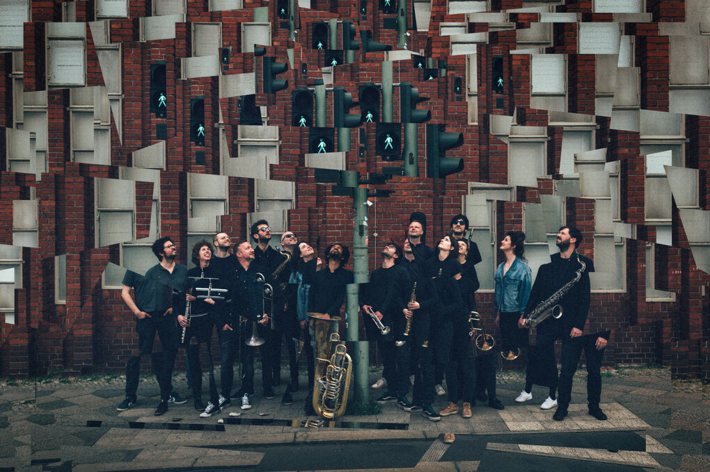 Eine Fotocollage des Magnetic Ghost Orchestra, die vor einem Backsteinbau mit grauen Fenstern unter einer Ampel stehen. Die Musiker:innen tragen Blasinstrumente. Duch einen Mosaikeffekt wiederholen und überlappen sich einzelne Bildelemente und Bildkanten.