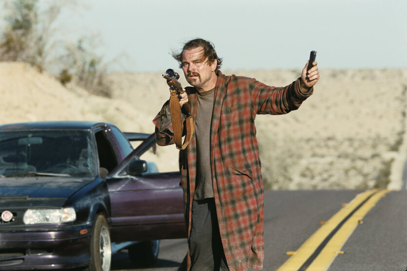 Bob (Leonardo DiCaprio) steht in Bademantel und mit Gewehr neben einem Auto auf der Landstraße.